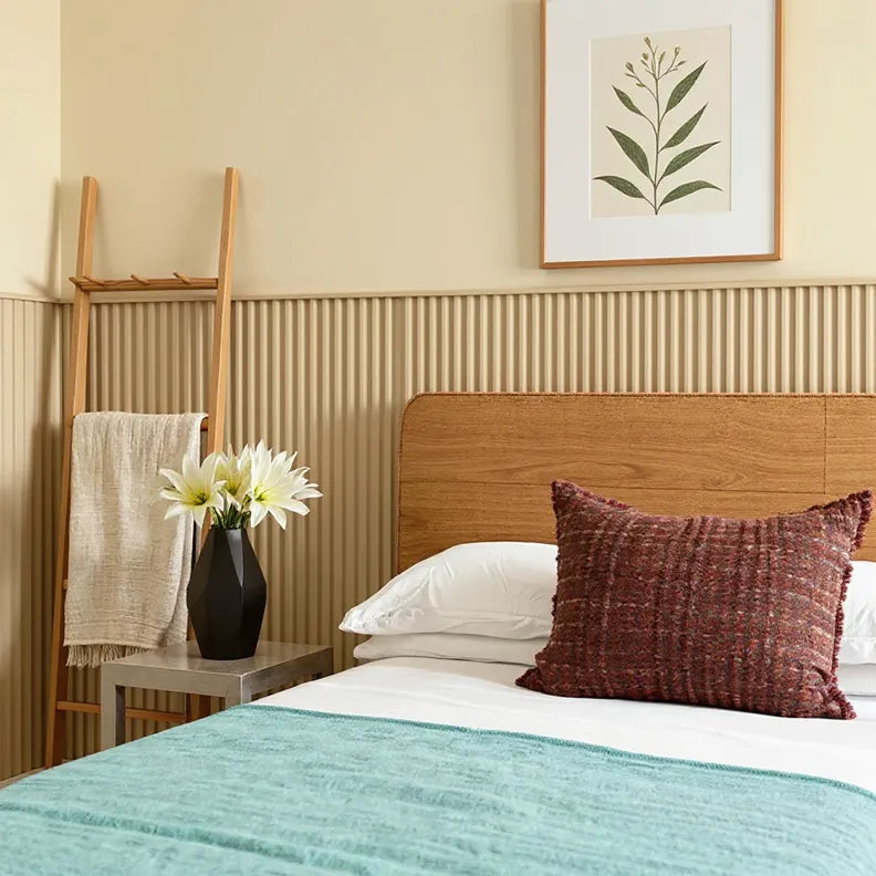 Modern bedroom interior featuring a wooden headboard bed with white bedding, teal blanket, and a textured maroon pillow. Behind the bed is a beige fluted wall panel accent. A black vase with white flowers sits on a small bedside table, and a wooden towel ladder leans against the wall. A framed botanical print hangs above the paneling.
